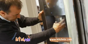 A man troubleshooting an ice maker, carefully inspecting the unit while defrosting built-up ice. He is using a hairdryer to melt the frost inside the machine, ensuring proper functionality. The ice maker is a countertop model with a transparent lid, showing partially frozen ice cubes inside.